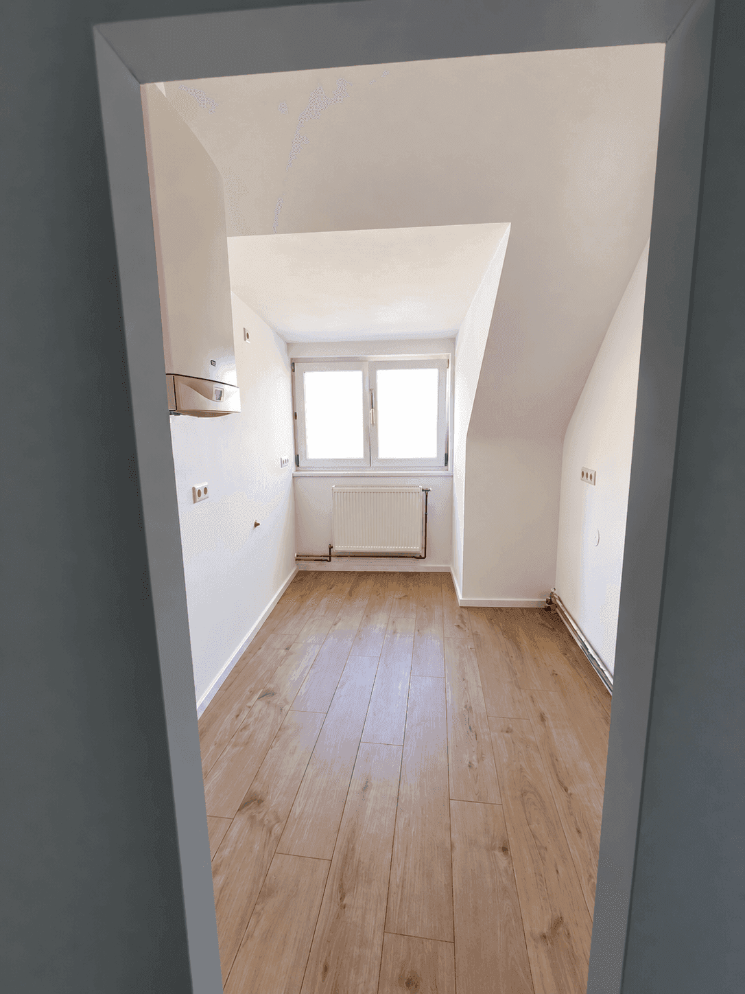 Leerer Küchenraum mit Dachschräge, Fenster und Anschlüssen für eine zukünftige Einbauküche.