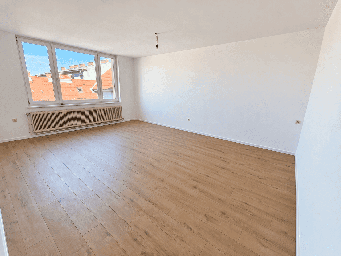 Helles, leeres Zimmer mit Laminatboden und mehreren Fenstern, die viel Tageslicht hereinlassen.