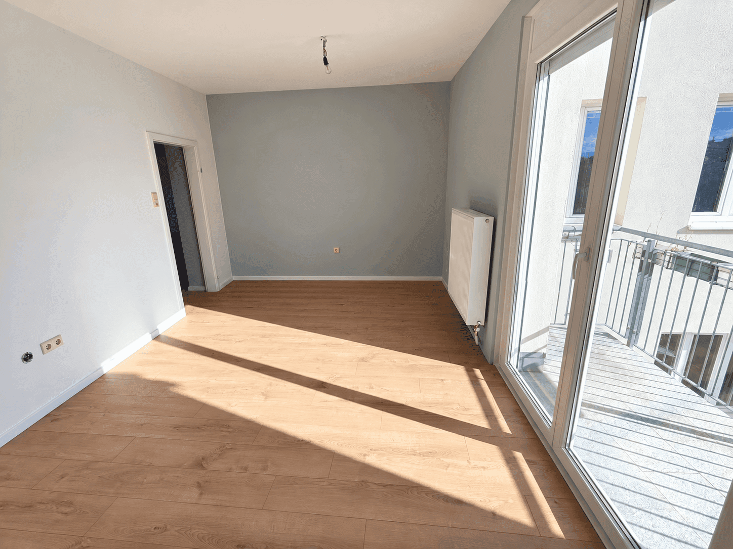 Leeres Zimmer mit hellem Laminatboden, hellblauen Wänden und Zugang zu einem kleinen Balkon.