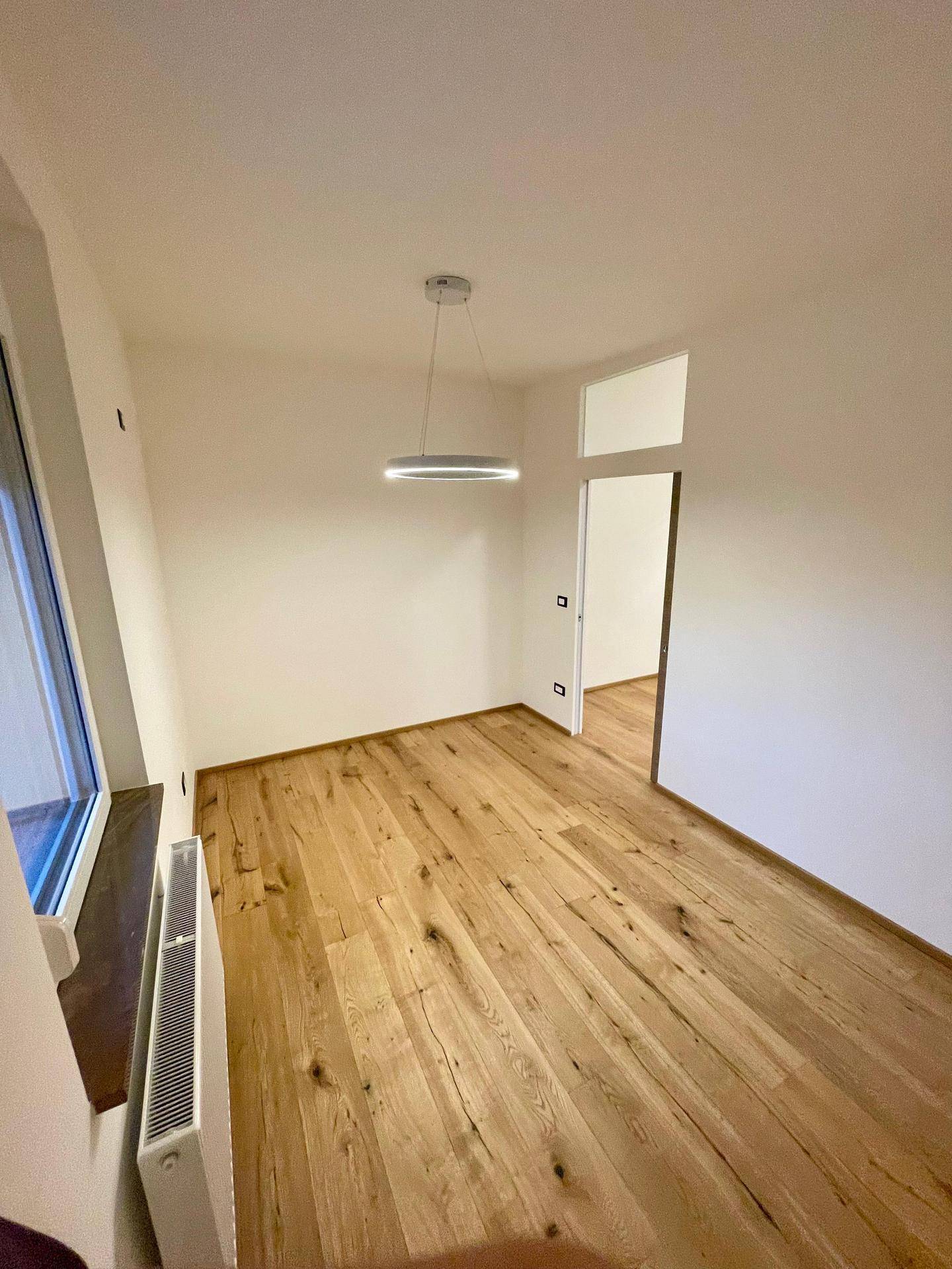 Leeres Zimmer mit Holzoptik-Fliesenboden, Heizkörper unter dem Fenster und moderner Hängelampe.