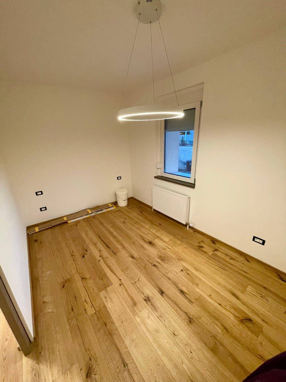 Leeres Schlafzimmer mit Holzoptik-Fliesenboden, großem Fenster und moderner Deckenleuchte.