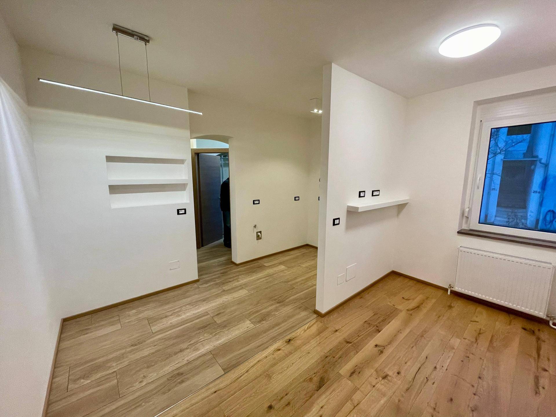 Heller Wohnbereich mit Holzoptik-Fliesenboden, integrierten Wandregalen und großem Fenster.