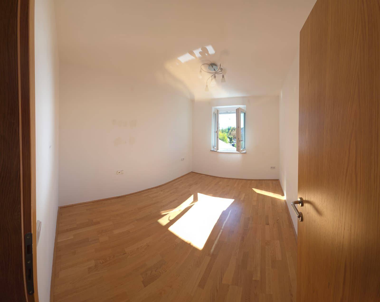 Helles, leeres Zimmer mit Holzboden und einem Fenster, das viel Sonnenlicht hereinlässt.