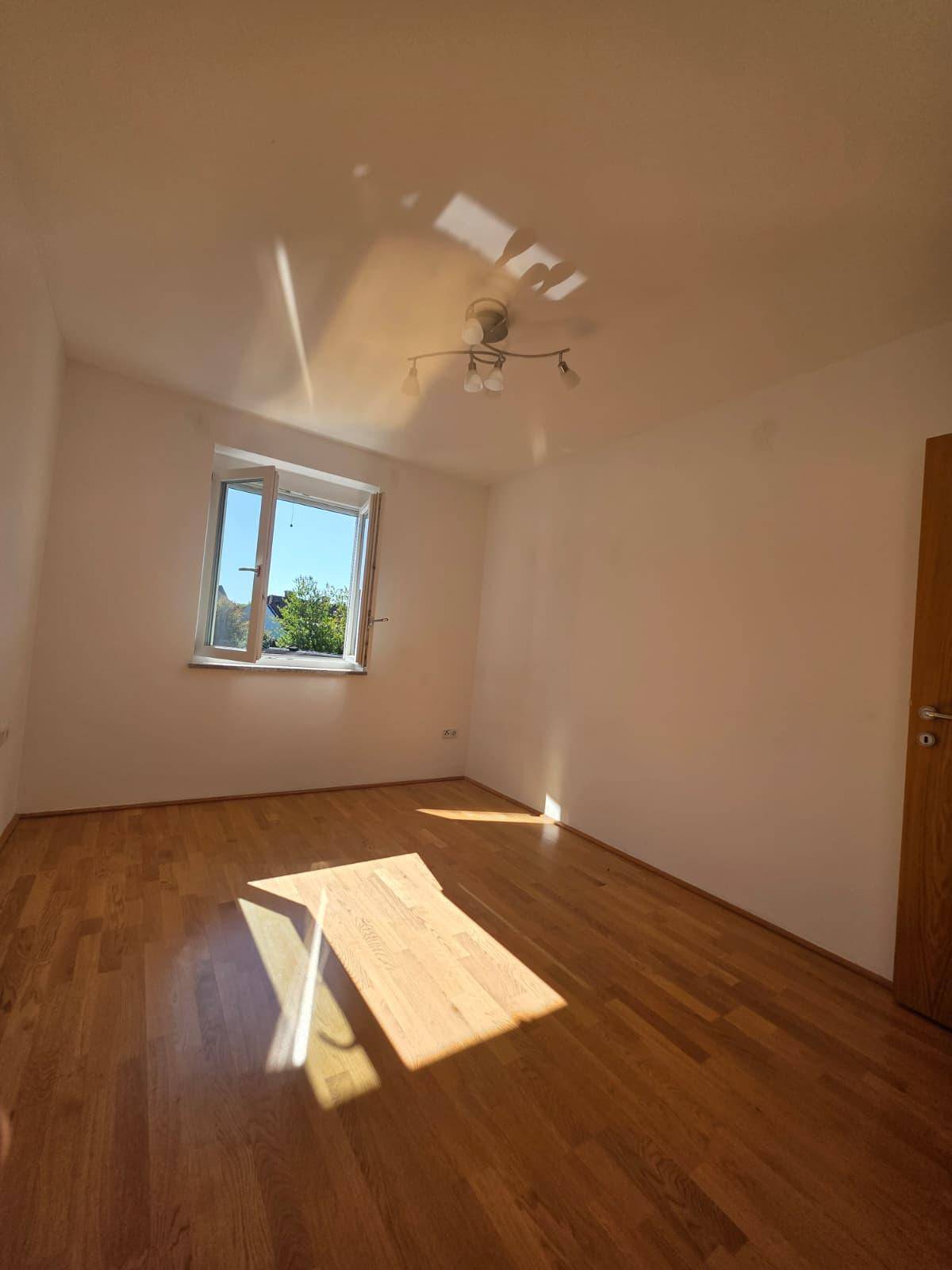 Geräumiges, leeres Zimmer mit Holzboden und einem Fenster, das viel Sonnenlicht hereinlässt.