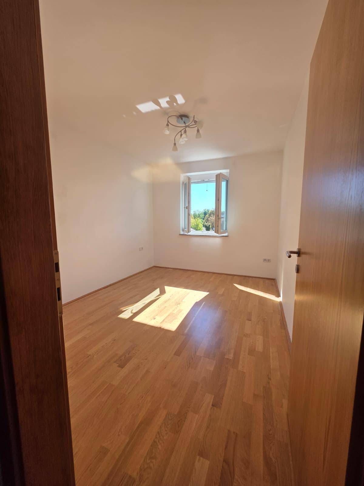 Helles, leeres Zimmer mit Holzboden und einem Fenster, das viel Sonnenlicht hereinlässt.