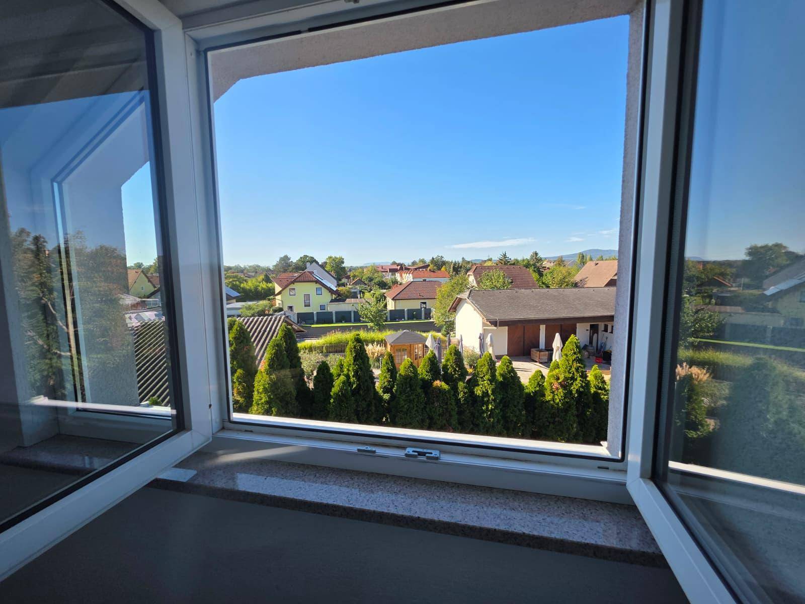 Weitblick aus dem Fenster über eine ruhige Wohnsiedlung mit viel Grün und blauen Himmel.