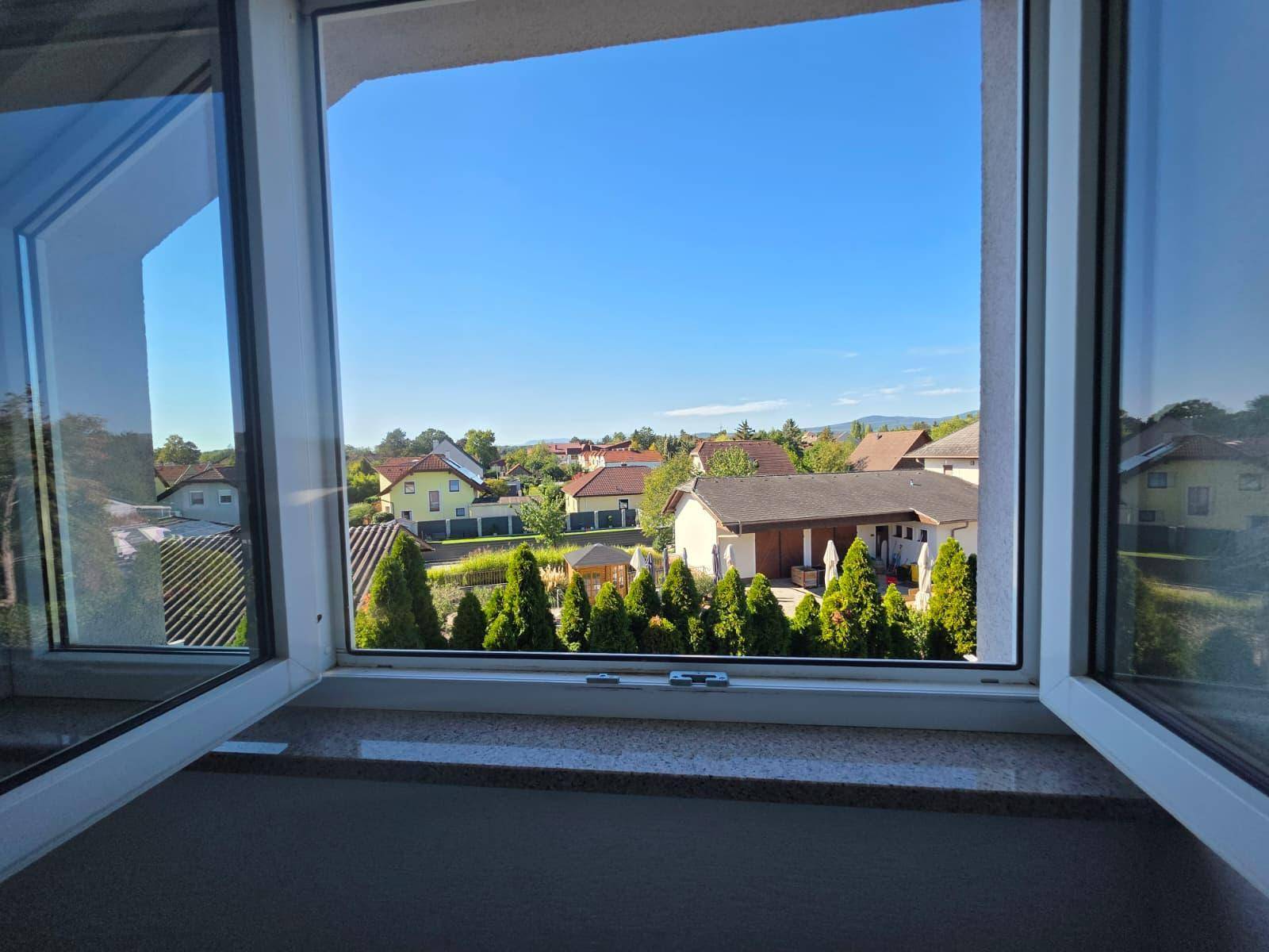 Weitblick aus dem Fenster über eine ruhige Wohnsiedlung mit viel Grün und blauen Himmel.