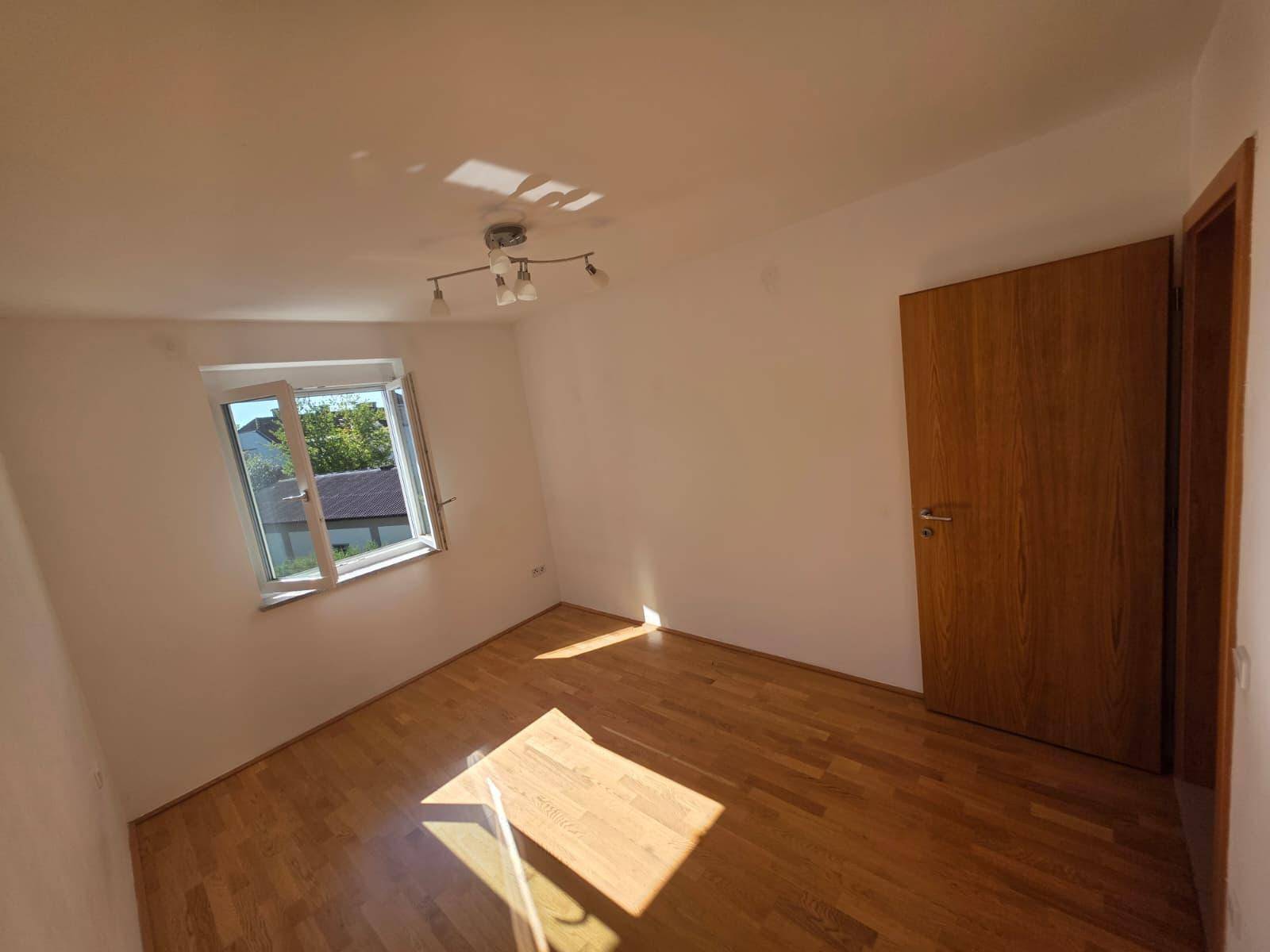 Leeres Zimmer mit Holzboden und einem Fenster, das Sonnenlicht auf den Boden wirft.
