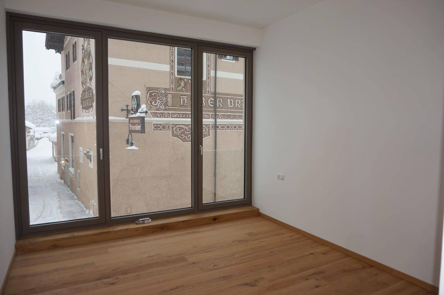 Helles Zimmer mit Holzboden und großen Fenstern mit Blick auf eine verschneite Straße und Gebäude.