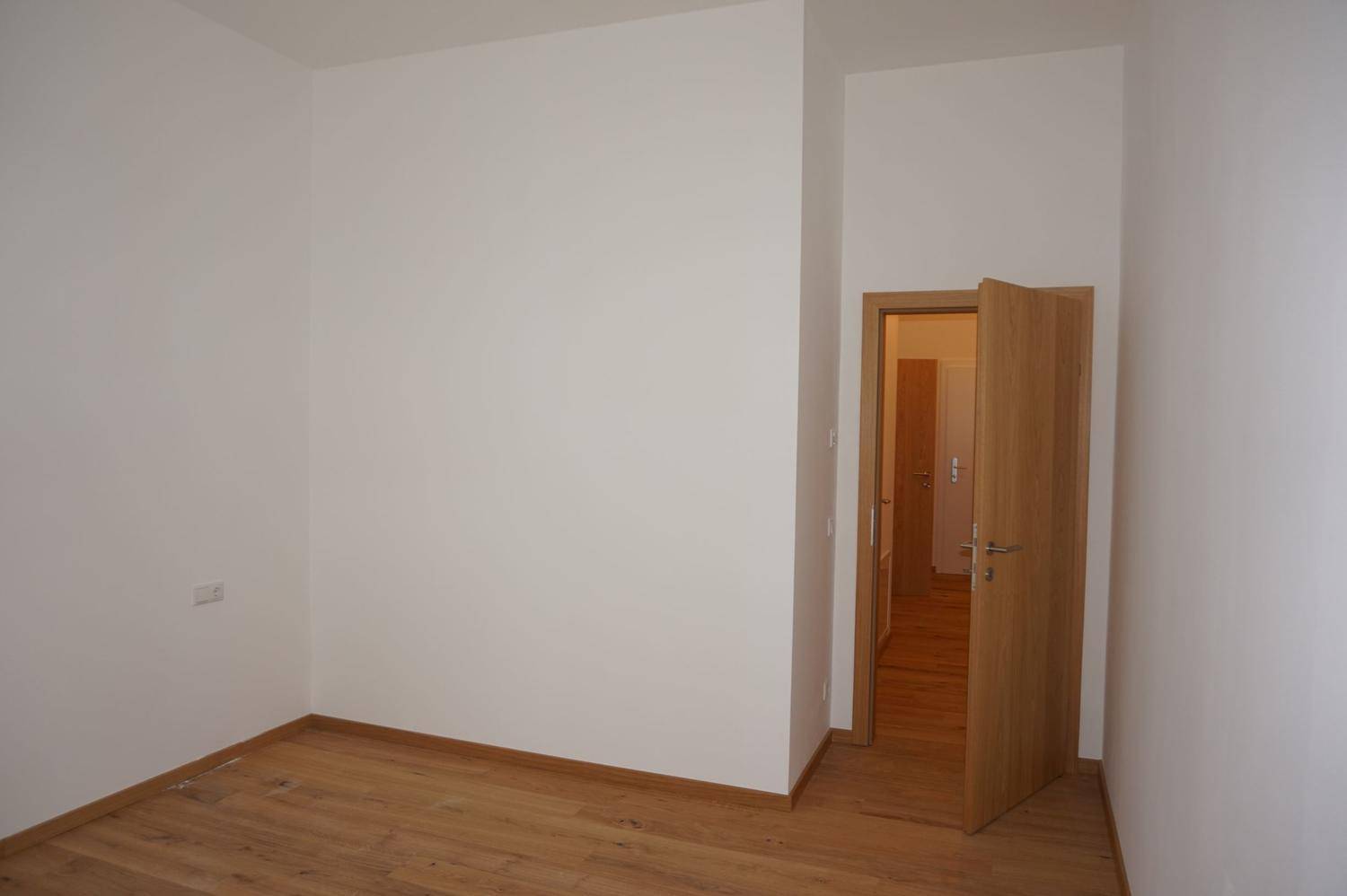 Leeres Zimmer mit Holzboden und weißen Wänden, eine offene Holztür führt in einen Flur.