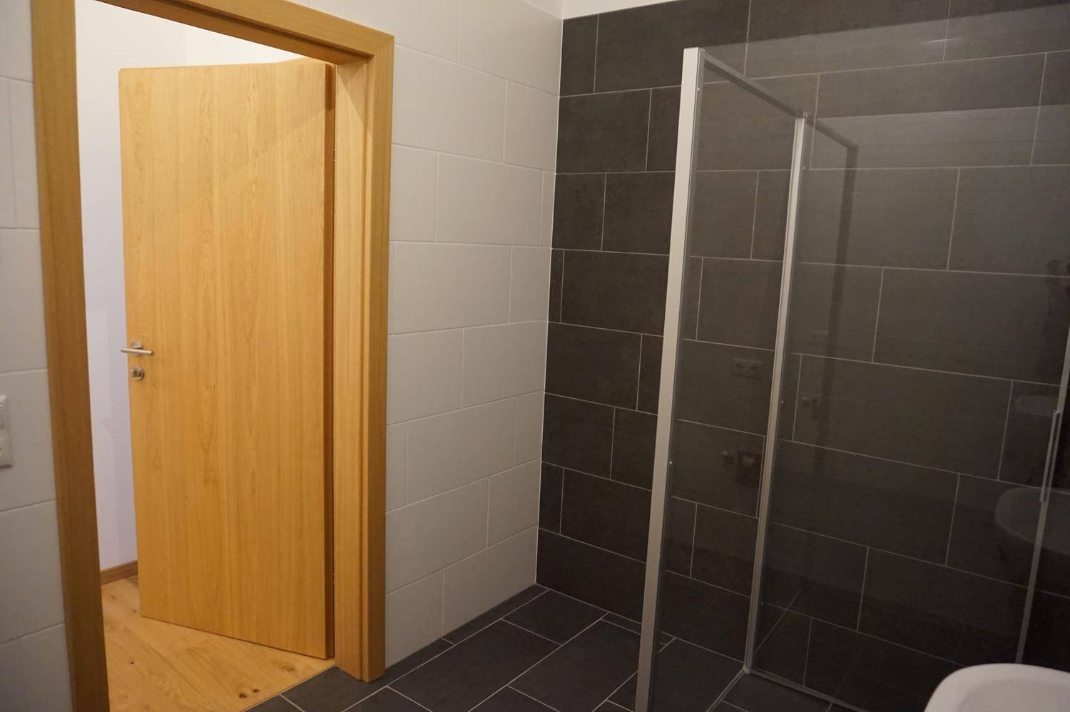 Badezimmer mit moderner Dusche und Holztür, die zu einem angrenzenden Raum mit Holzboden führt.