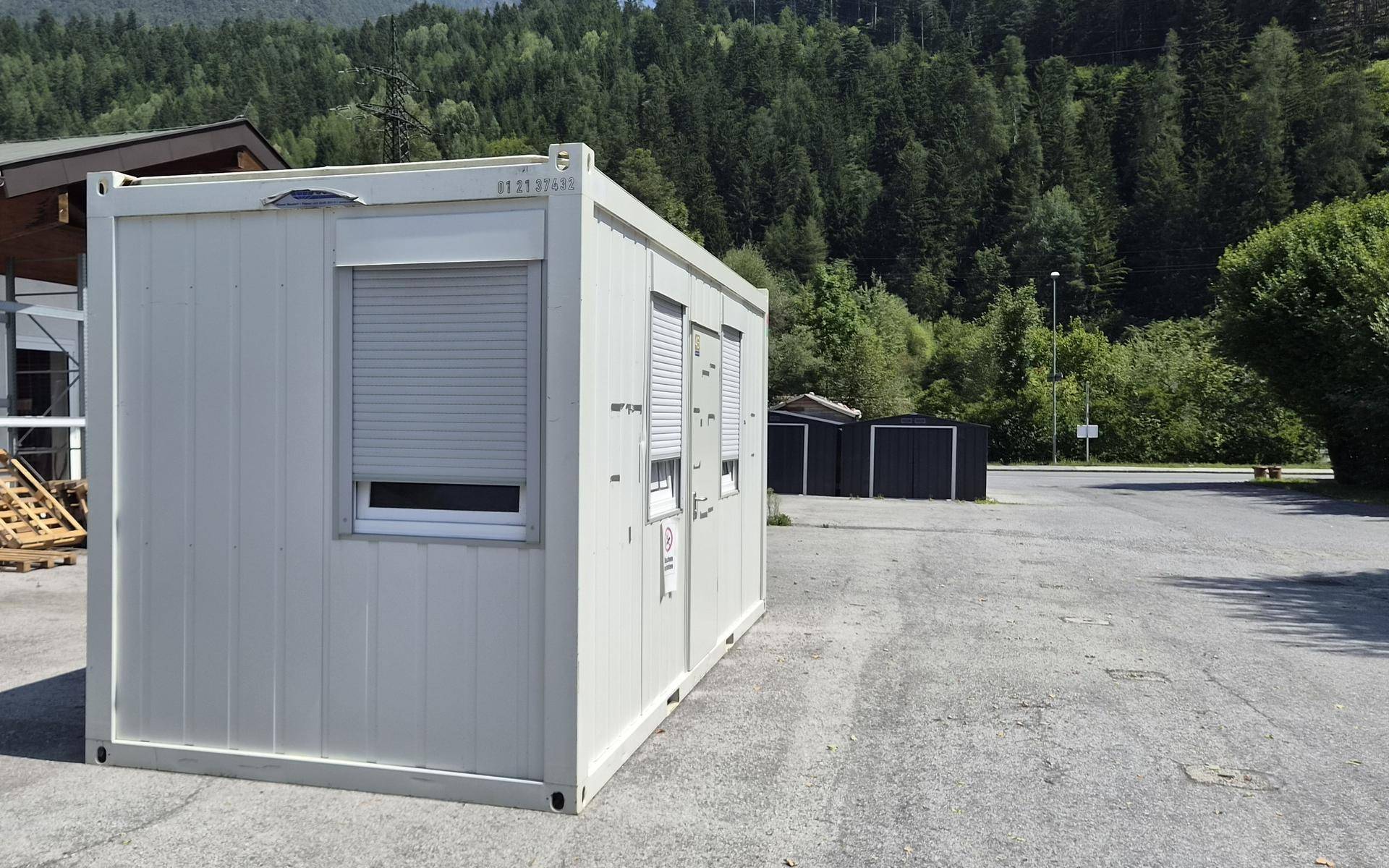 Seitenansicht des weißen Bürocontainers auf einem Parkplatz, umgeben von Bäumen und Bergen im Hintergrund.