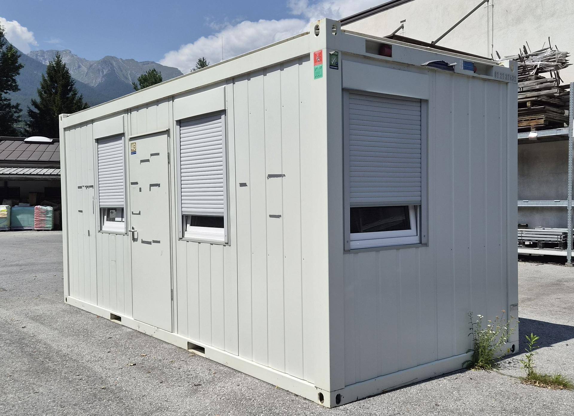 Schräge Ansicht des weißen Bürocontainers mit Rollläden, im Hintergrund sind Berge und ein Lagerbereich zu sehen.
