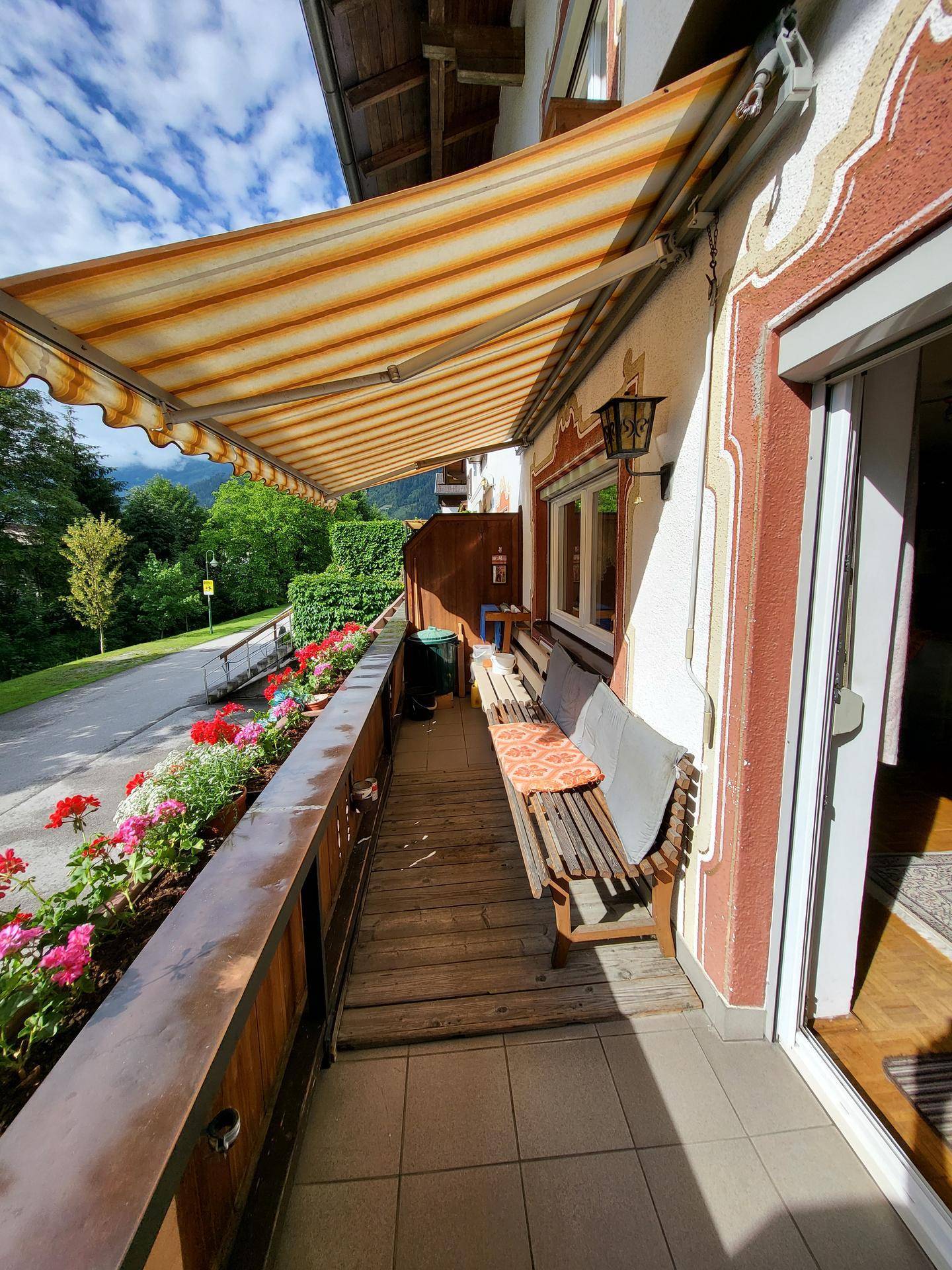 Sonniger Balkon mit gestreifter Markise, Holzboden und Blumenkästen mit Bergblick.