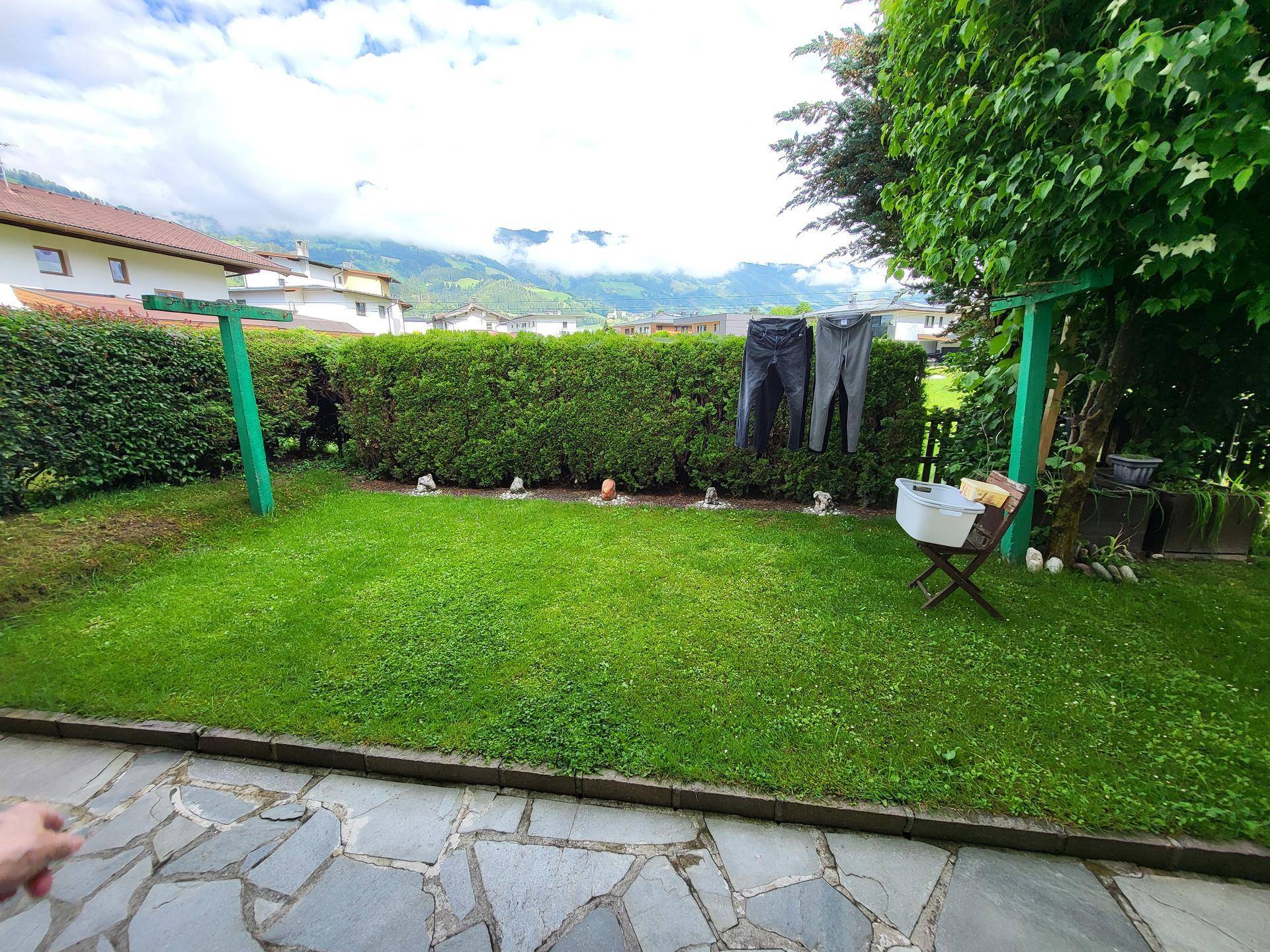 Gepflegter Garten mit Rasenfläche, Hecke und Wäscheleine, umgeben von Bergen.