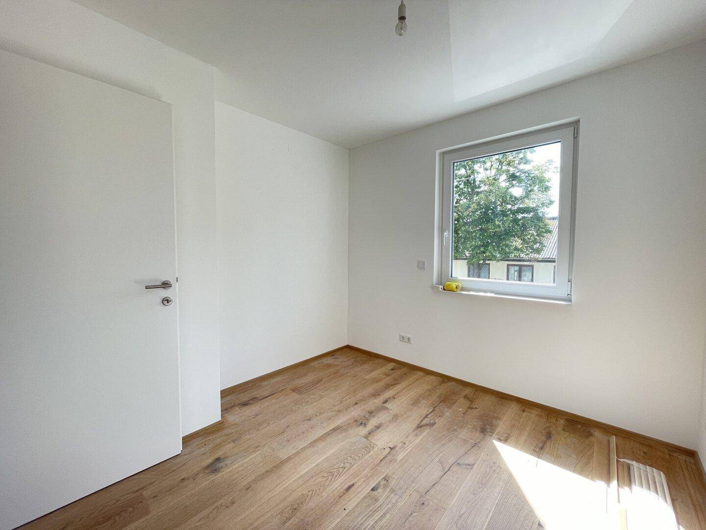Lichtdurchflutetes Zimmer mit Parkettboden, weißen Wänden und großem Fenster.