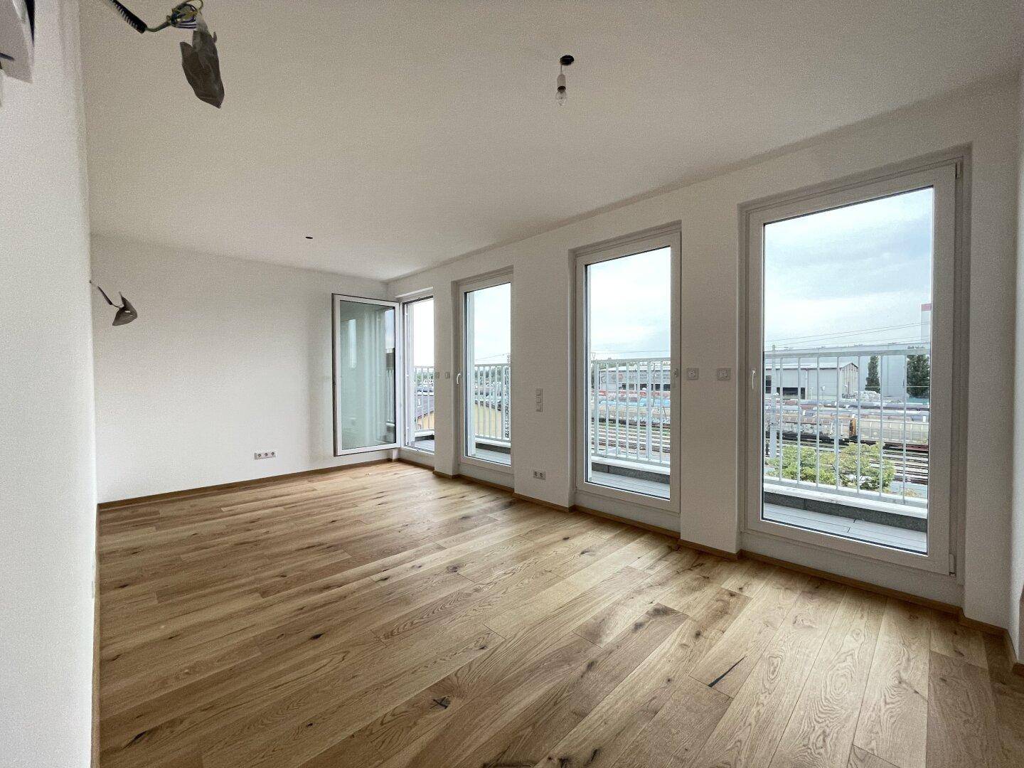 Geräumiges Wohnzimmer mit Holzboden und mehreren Fenstertüren, die auf einen Balkon führen.
