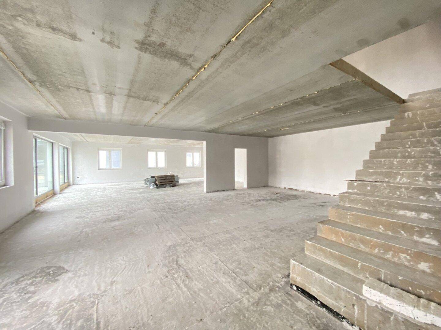 Großer, offener Raum im Rohbau mit Betondecke, Treppe und mehreren Fenstern, ideal für Büros.