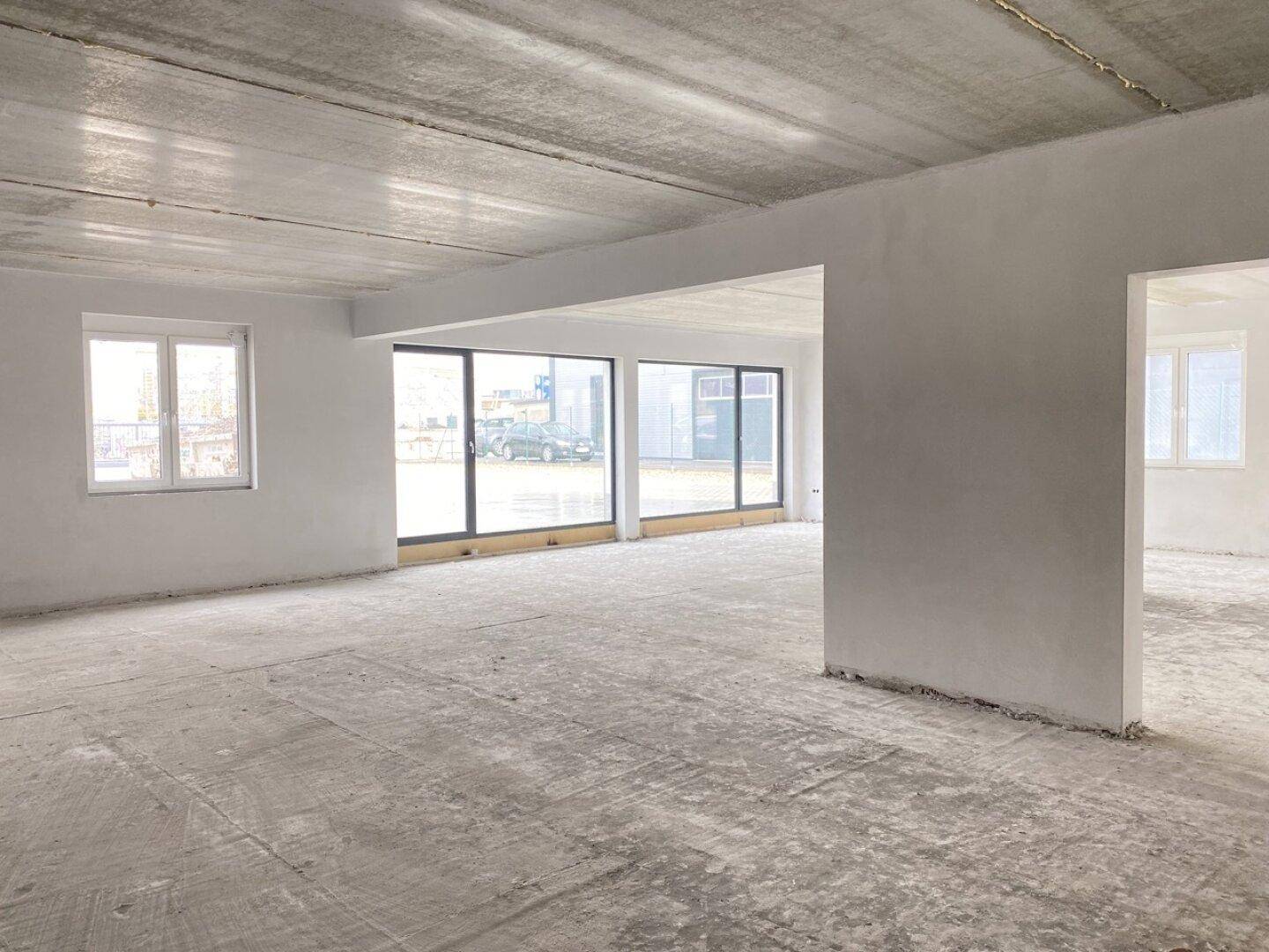 Offener Büroraum im Rohbau mit großen Fensterfronten und einer Betondecke, viel Tageslicht.