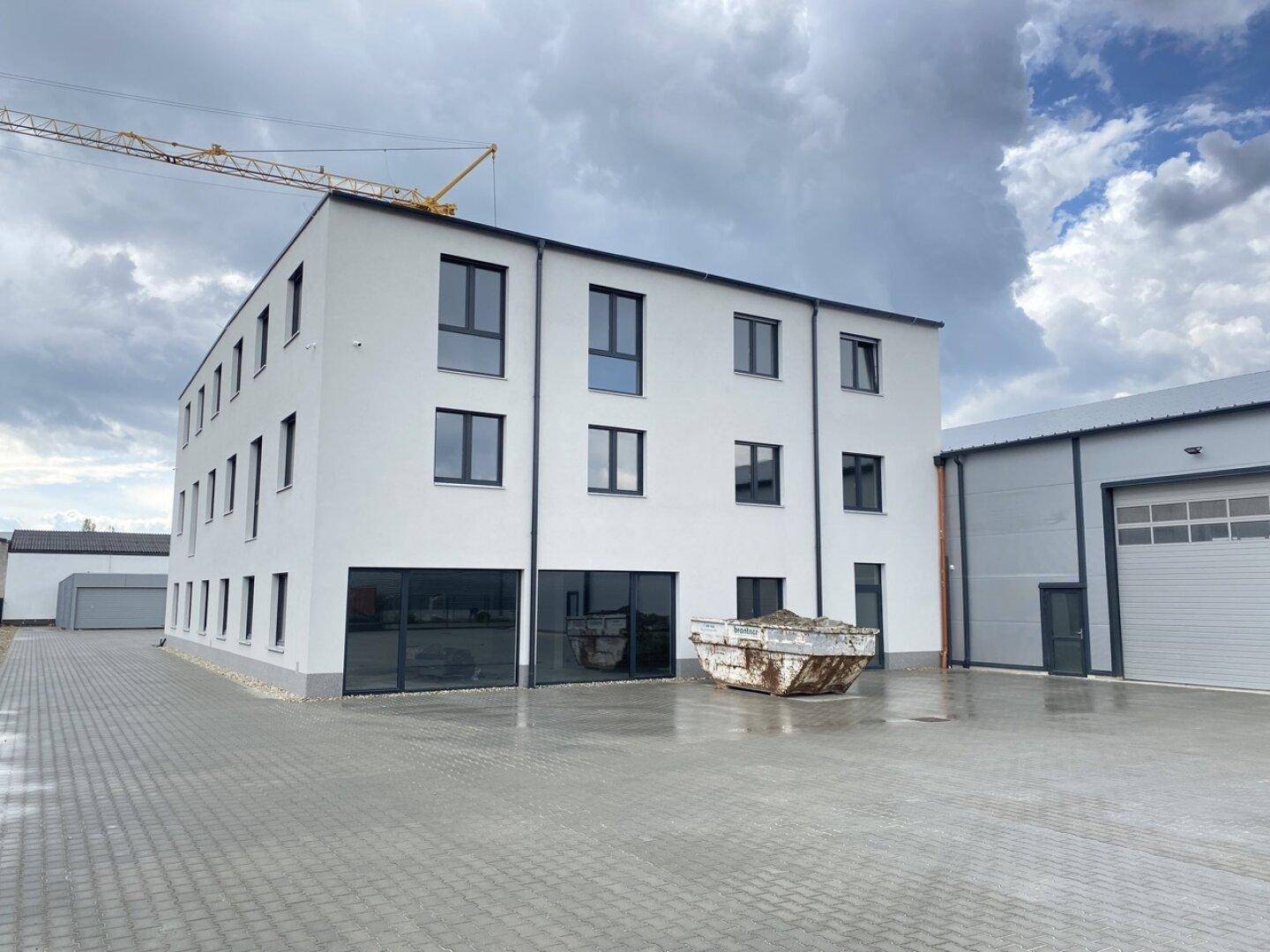 Modernes Bürogebäude und Lagerhalle mit gepflastertem Vorplatz unter bewölktem Himmel.
