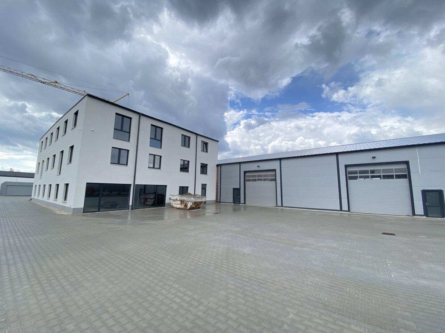 Gesamtansicht eines neuen Bürogebäudes und einer Lagerhalle mit großem Vorplatz und Kran im Hintergrund.