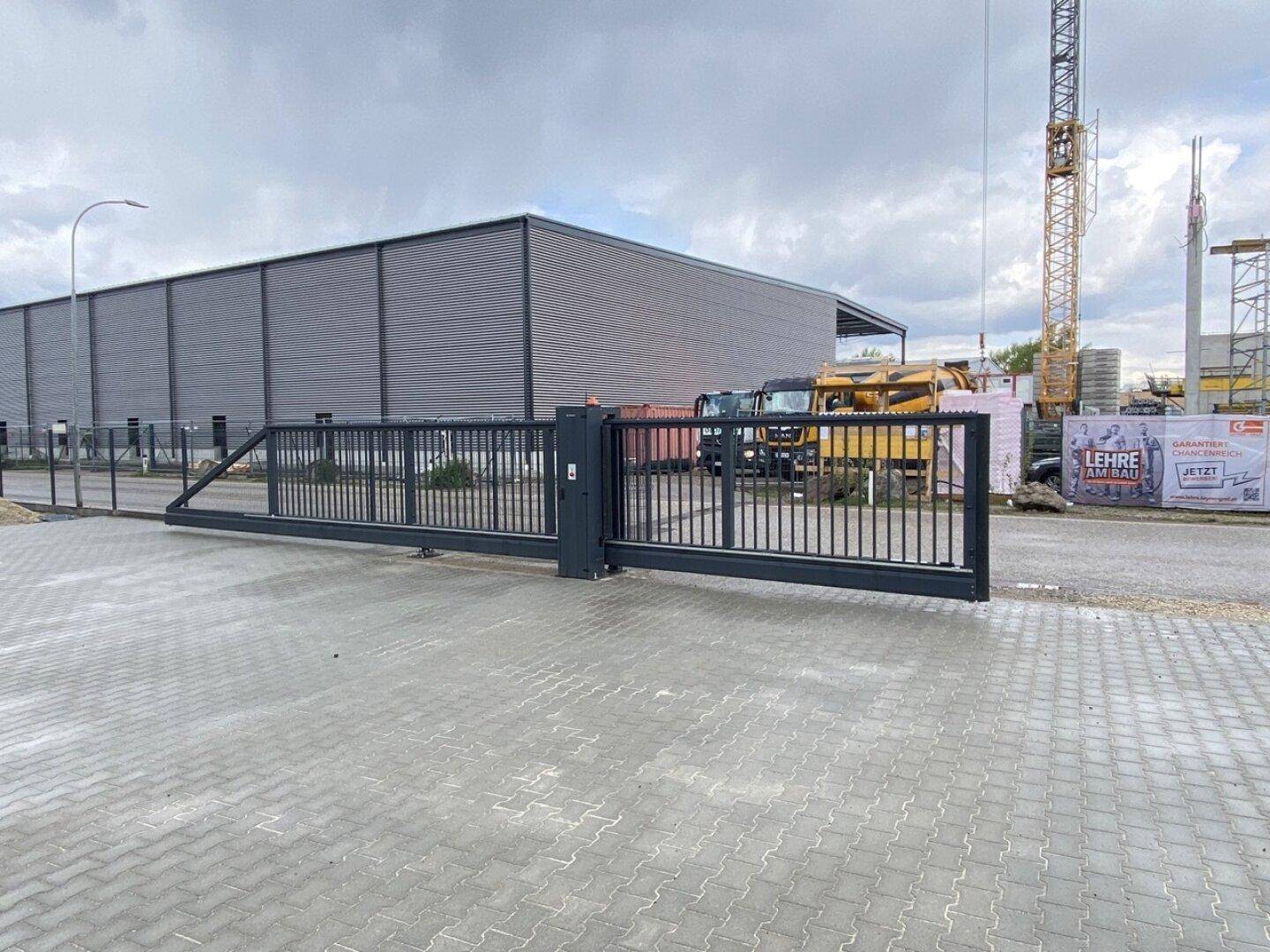 Zufahrt zu einem Industriegelände mit Schiebetor und gepflastertem Bereich, Baustelle im Hintergrund.