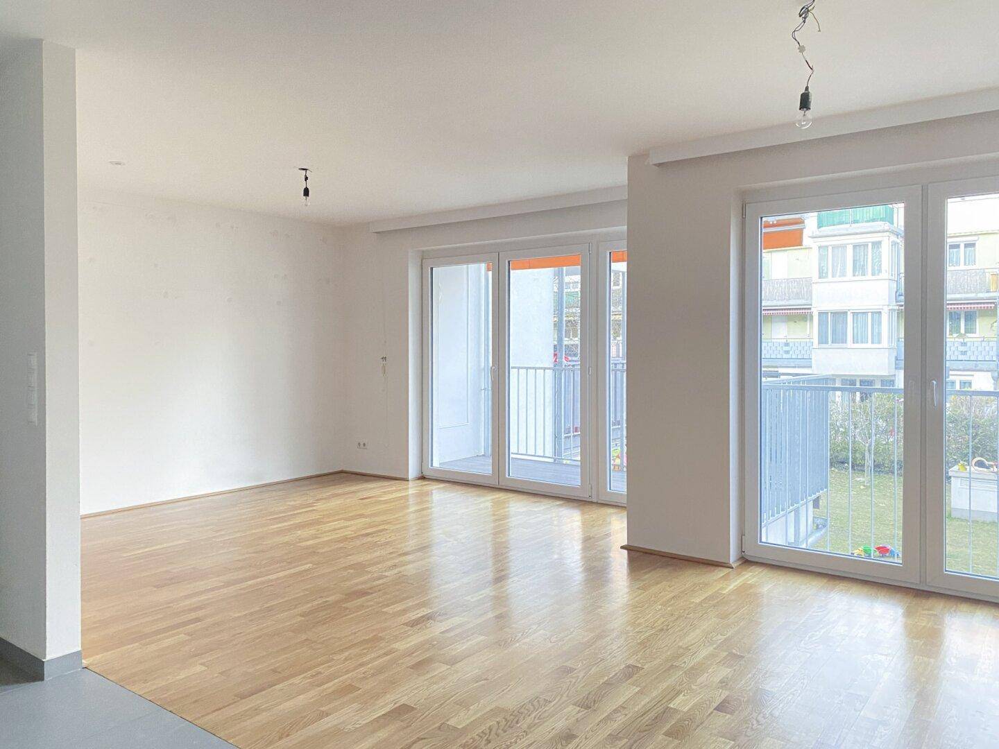 Geräumiger Wohnbereich mit hellem Parkettboden und großen Fenstern, die viel Tageslicht hereinlassen.
