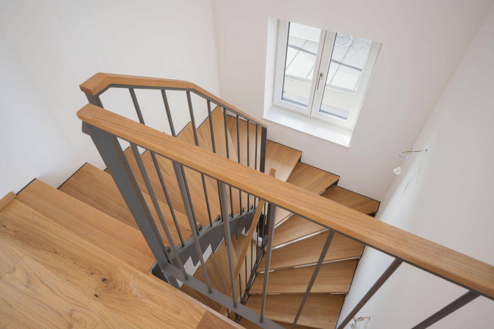 Moderne Holztreppe mit Metallgeländer und Fenster, die viel Licht hereinlässt.