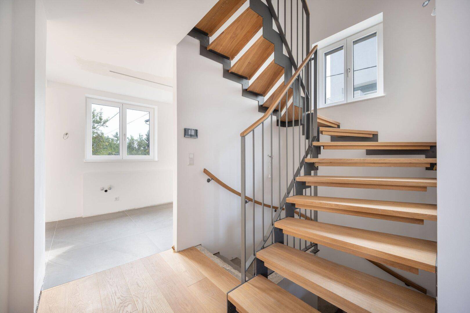 Offene Holztreppe mit Metallgeländer in einem hellen Eingangsbereich mit Fenstern.