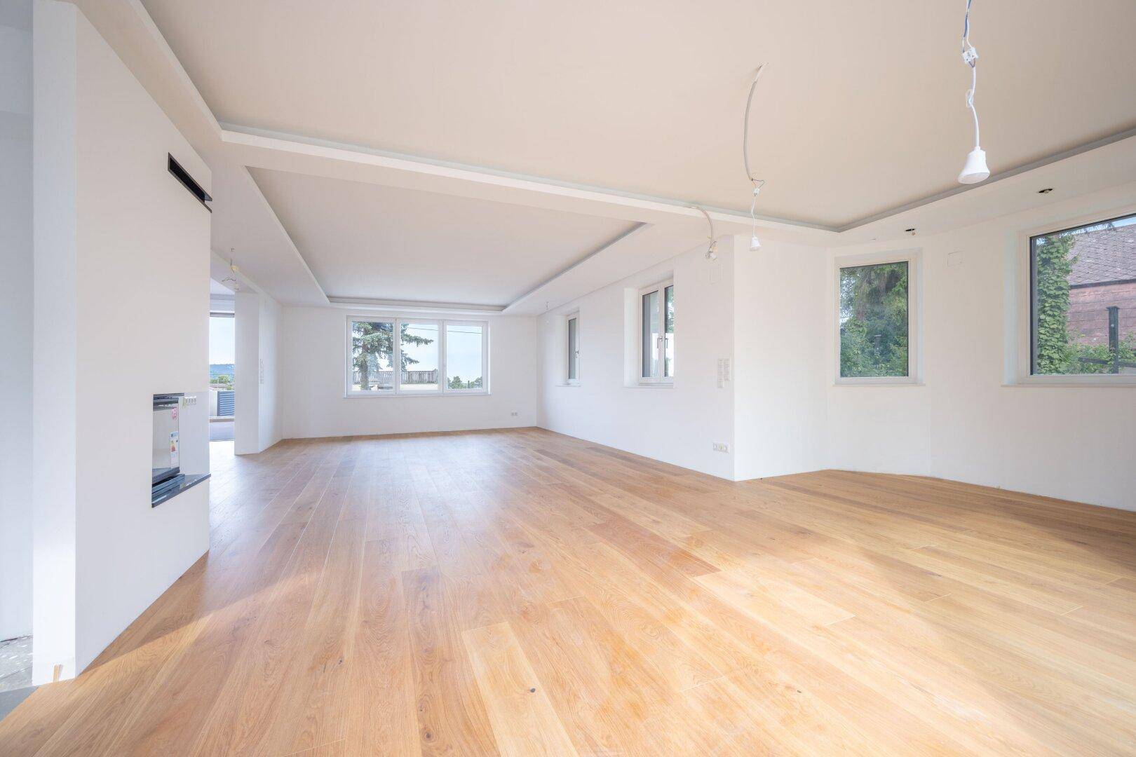 Geräumiger Wohnbereich mit Holzboden, Kamin und großen Fenstern, die viel Licht hereinlassen.