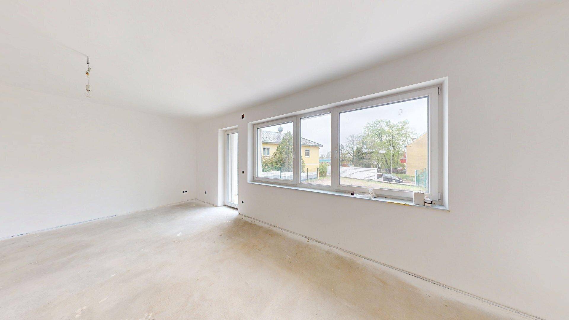 Geräumiges Wohnzimmer im Rohbau mit großen Fenstern, die viel Tageslicht hereinlassen.