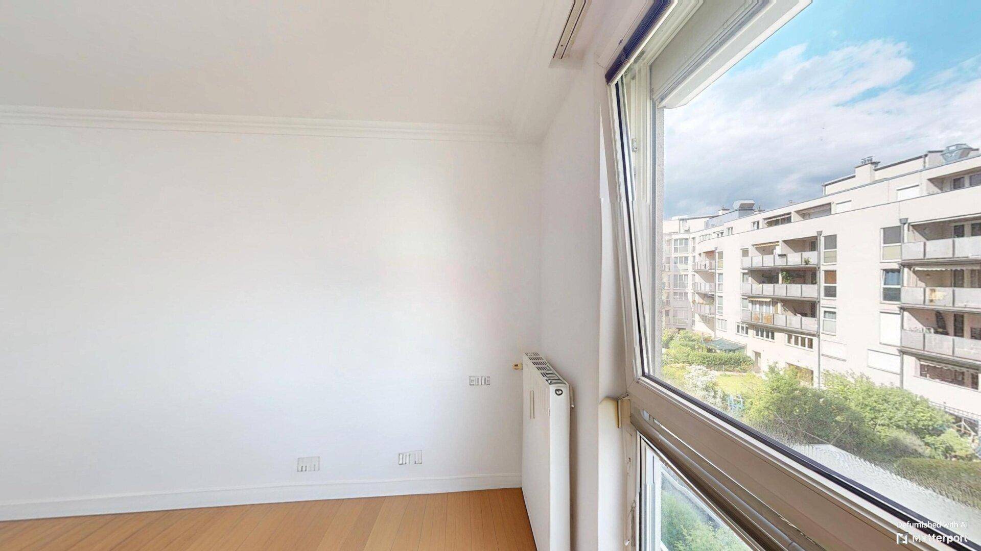 Leerer Raum mit Parkettboden, weißen Wänden und einem Fenster mit Blick auf die umliegenden Gebäude.