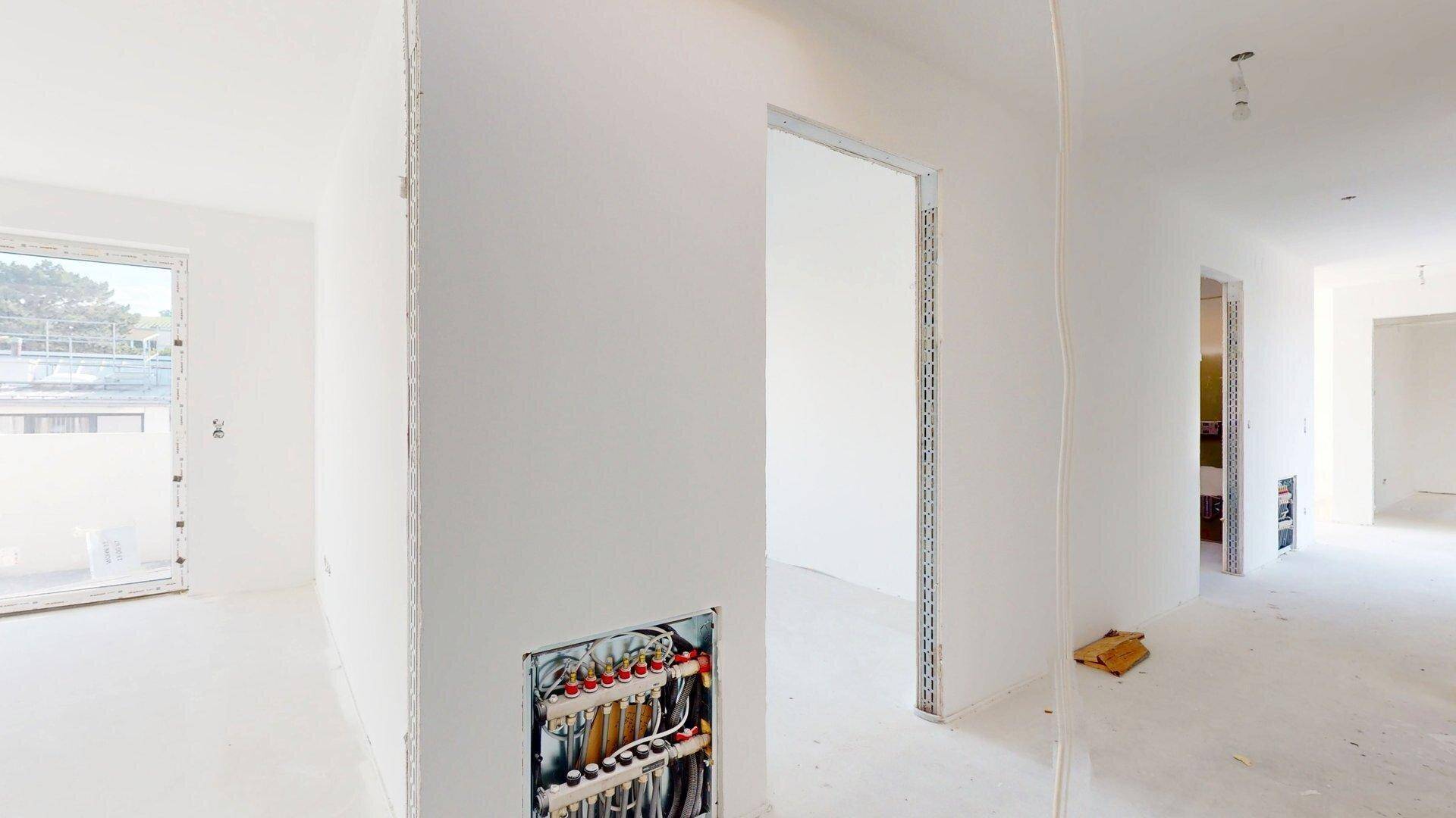 Flurbereich im Rohbau mit sichtbaren Installationen und offenen Türrahmen zu angrenzenden Räumen.