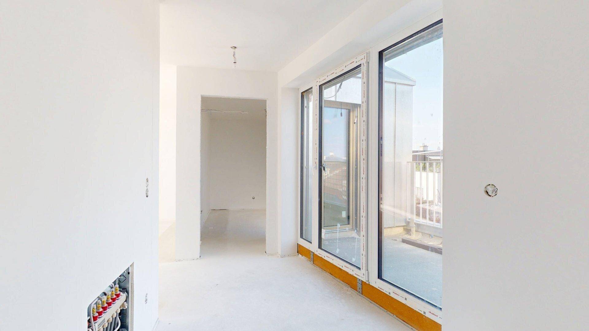 Unmöblierter Raum im Rohbau mit Fenster und sichtbaren Heizungsinstallationen an der Wand.