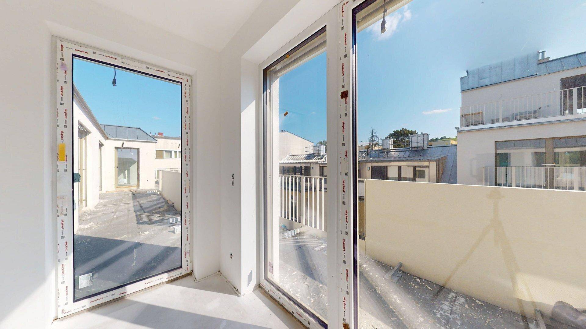 Blick aus dem Fenster auf eine unmöblierte Terrasse mit umliegenden Gebäuden unter blauem Himmel.