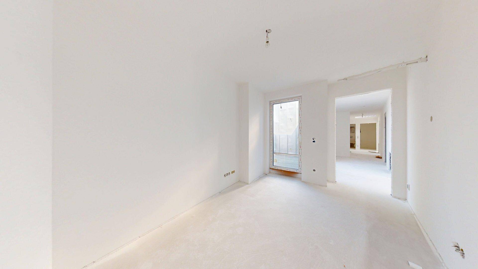 Unmöblierter Raum im Rohbau mit Fenster und offenem Zugang zu einem weiteren Bereich.