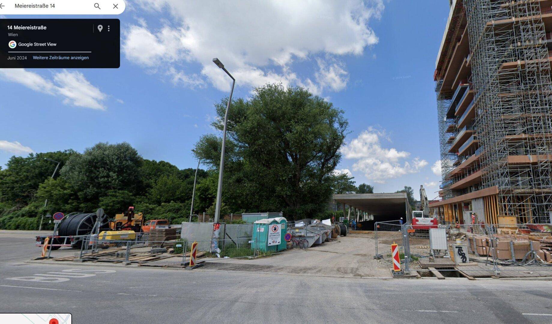 Straßenansicht einer Baustelle mit einem im Bau befindlichen Gebäude und umliegender Vegetation.