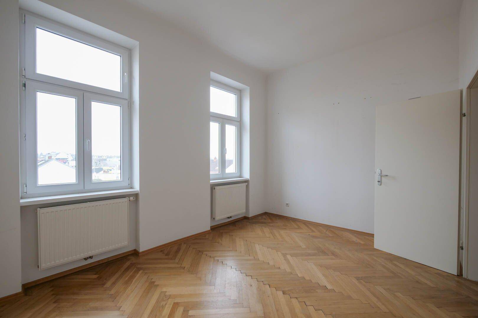 Helles Zimmer mit Fischgrätparkett und Fenstern, die den Raum mit Tageslicht durchfluten.