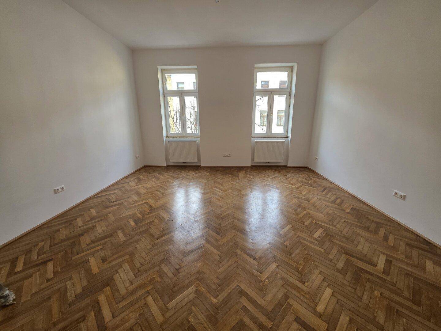Geräumiges Zimmer mit klassischem Fischgrätparkett und zwei großen Fenstern.