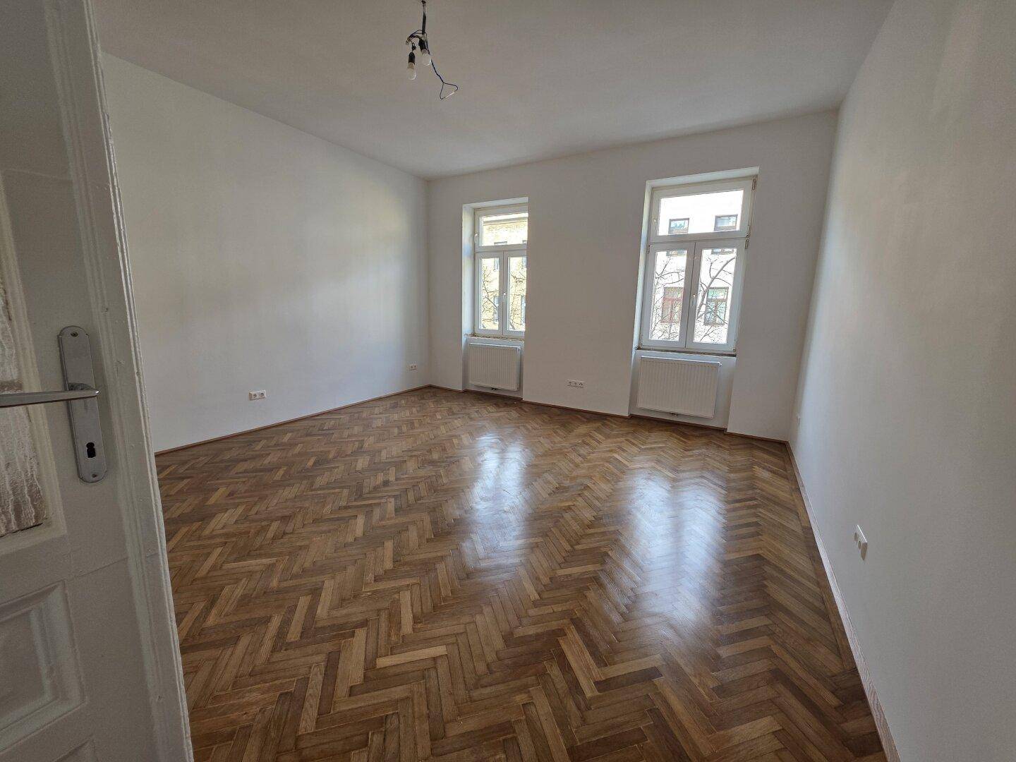 Leeres Zimmer mit glänzendem Fischgrätparkett und zwei Fenstern, bereit zur Einrichtung.