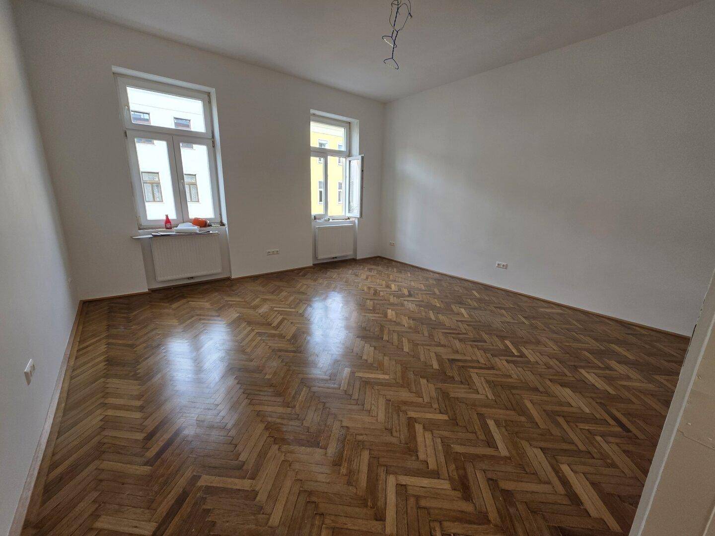 Helles Zimmer mit Fischgrätparkett und zwei Fenstern, die viel Tageslicht hereinlassen.