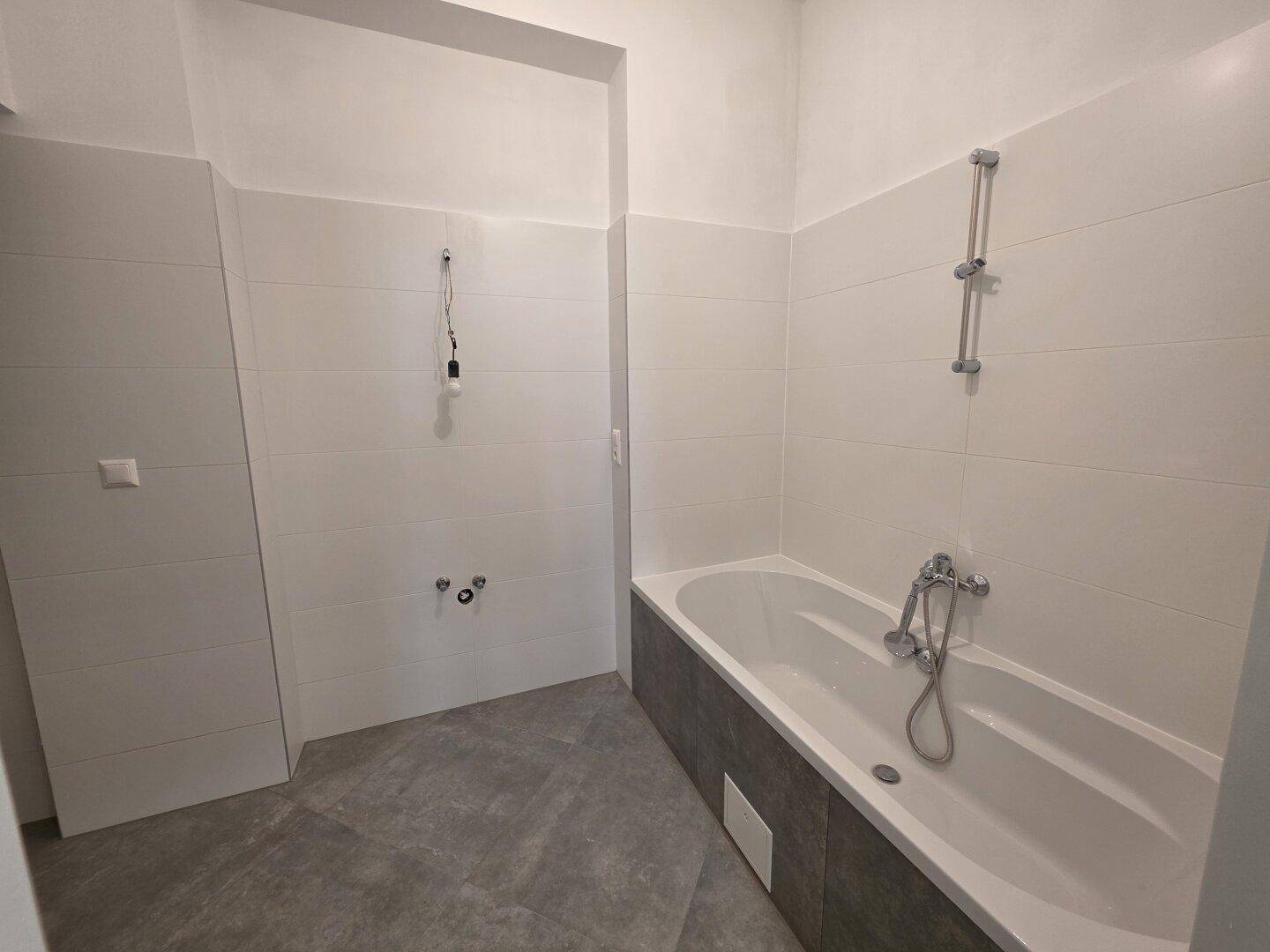 Modernes Badezimmer mit Badewanne und grauen Fliesen, bereit für die Installation der Armaturen.