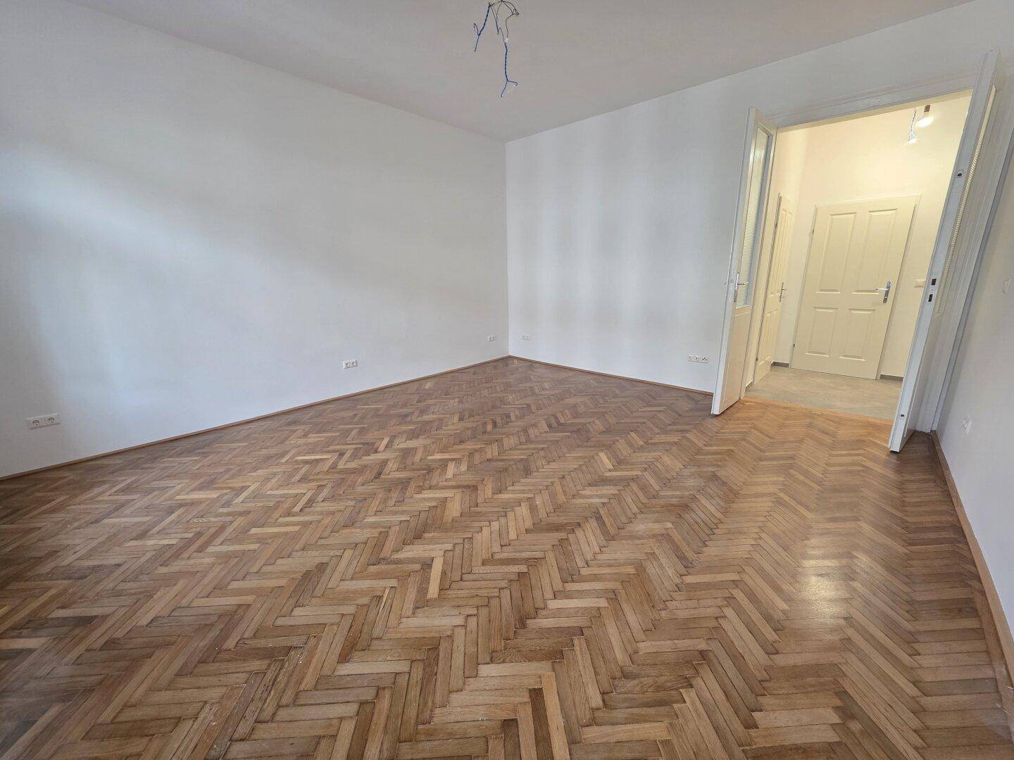 Geräumiges Zimmer mit Fischgrätparkett und einer Tür, die in den Flur führt.