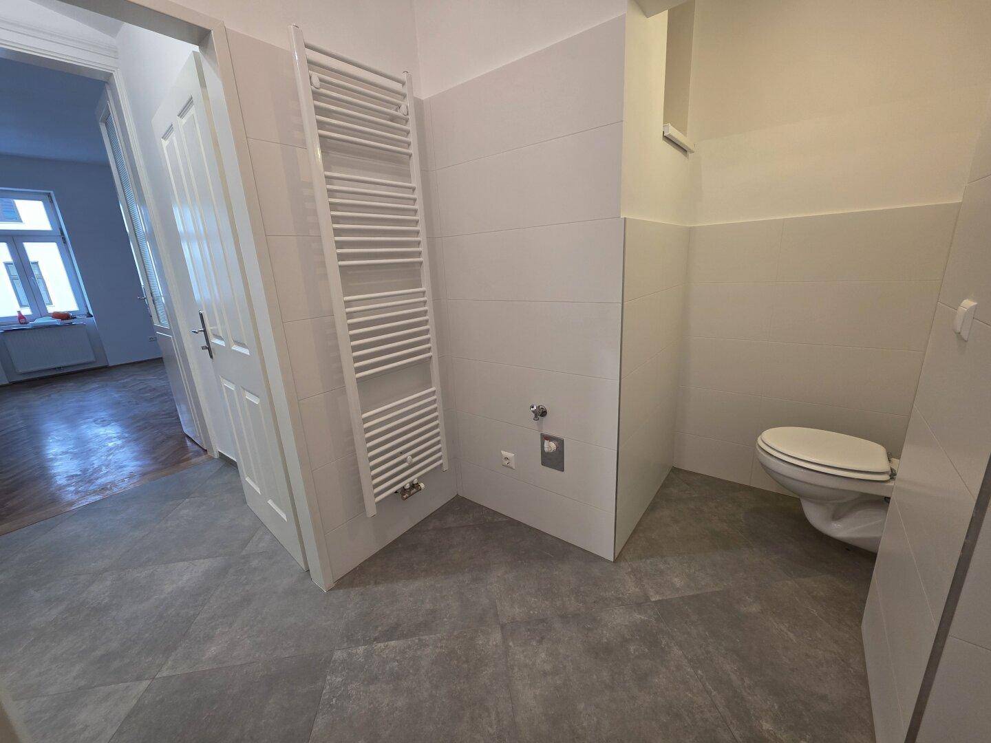 Badezimmer mit Wand-WC, beheiztem Handtuchhalter und grauen Bodenfliesen.