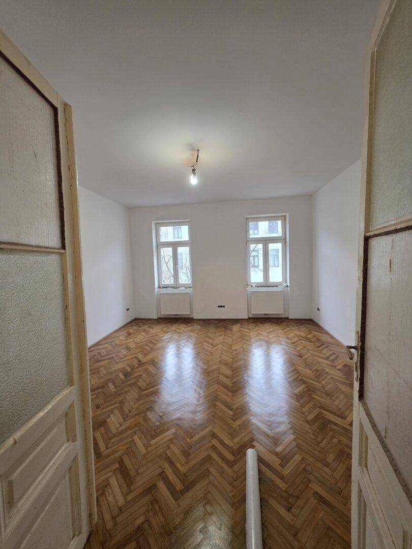 Großes Zimmer mit Fischgrätparkett und zwei Fenstern, die den Raum mit Licht füllen.