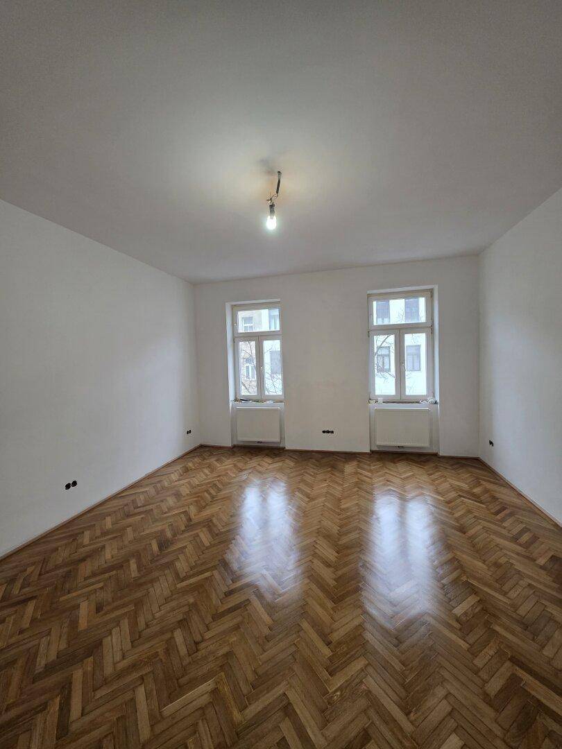 Geräumiges Zimmer mit Fischgrätparkett und zwei Fenstern, die für viel Helligkeit sorgen.