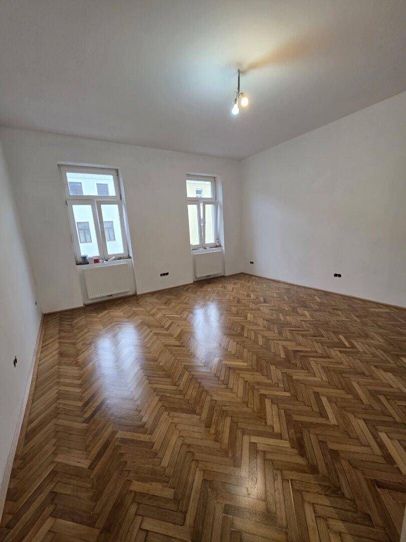 Großes Zimmer mit glänzendem Fischgrätparkett und zwei Fenstern, die den Raum erhellen.
