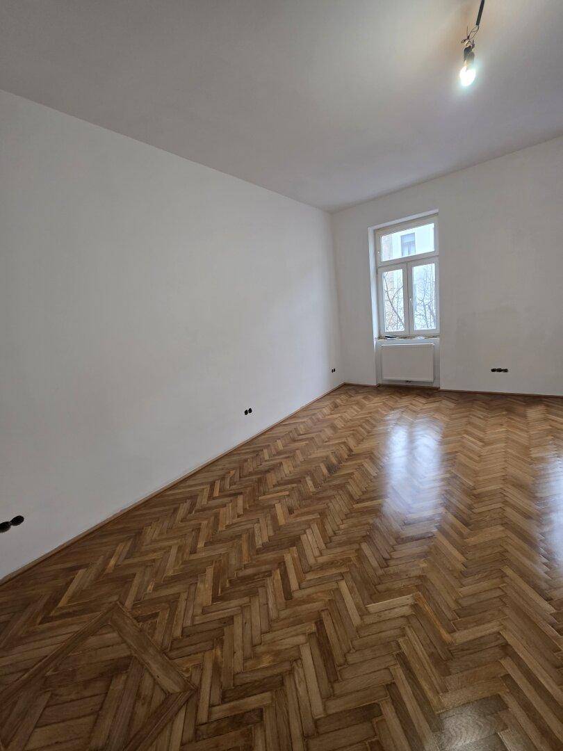Helles Zimmer mit Fischgrätparkett und einem großen Fenster, das den Raum erhellt.