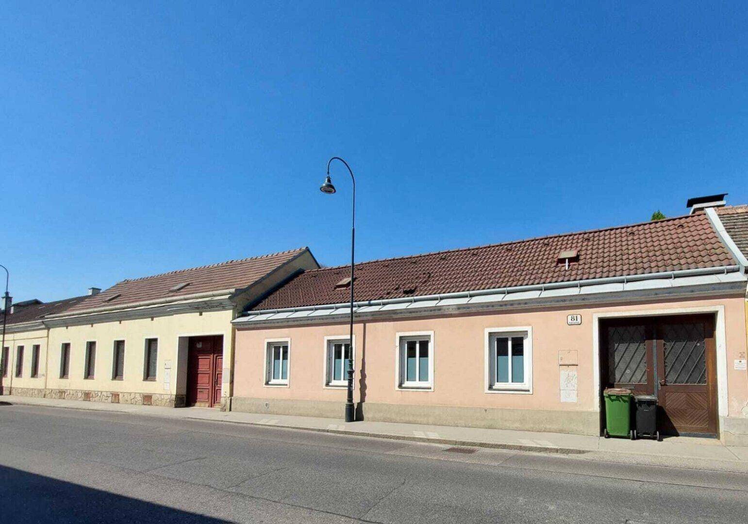 Straßenansicht mit zwei farbigen Altbauten, Ziegeldächern und einer Straßenlaterne unter blauem Himmel.