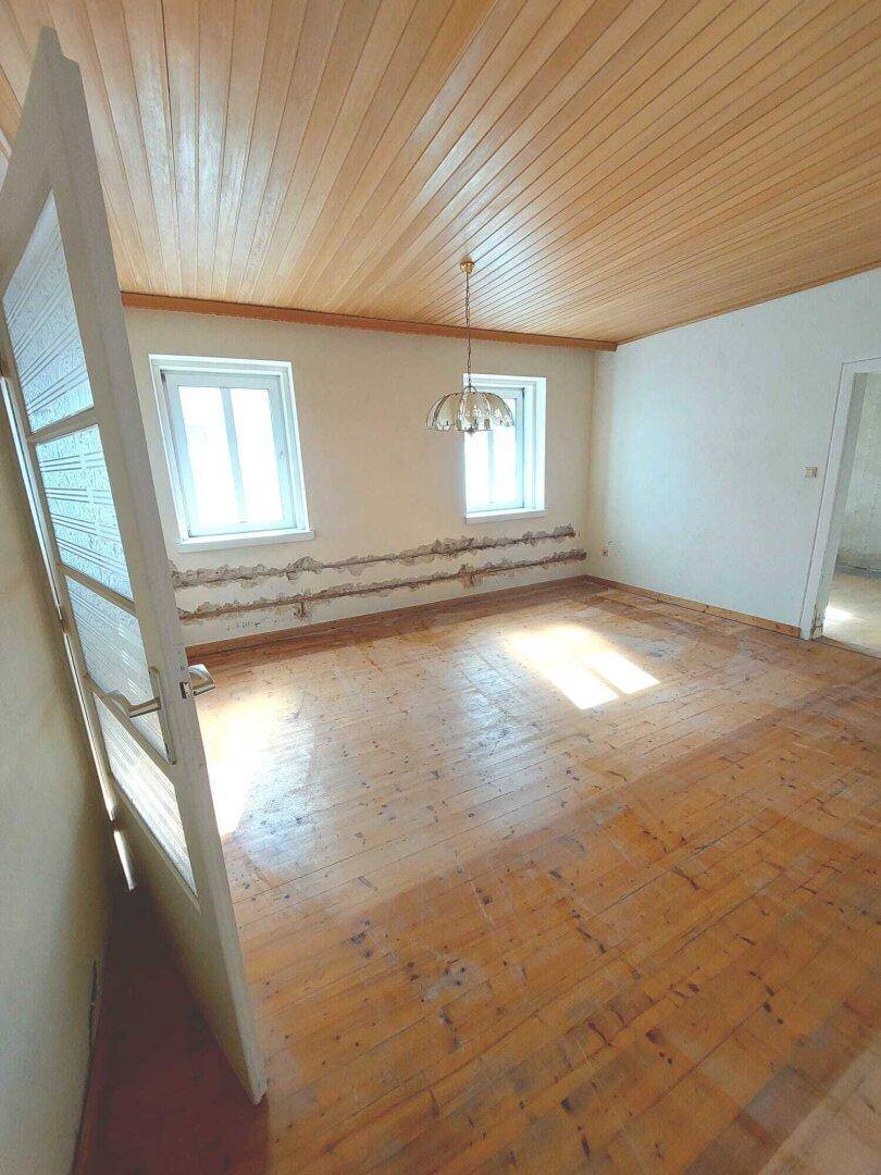 Helles Zimmer mit Holzdielenboden, Holzdecke und zwei Fenstern, bereit zur Renovierung.